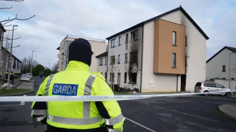 A uniformed garda (Irish police) officer stands guard at a white cordon outside a house in Creston Avenue, Finglas, following an arson attack.   The officer, wearing a yellow high-viz jacket and a black snood, has their back to the camera.  They are looking towards a cream-coloured three-store building which shows signs of smoke-damage on all three floors.  Burnt furniture and belongings are scattered in the front garden.