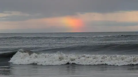 BBC Weather Watchers/Kathryn The sea which has looks fairly calm and grey. There is a small wave in the foreground with white water in the front. The sky is cloudy and grey but in the centre of the photo there is a streak of a rainbow with red, orange and green colours.
