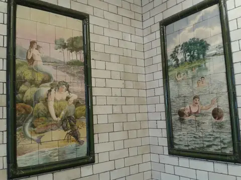 Empty Images Two white-tiled walls with a picture framed on each one. The left-hand tiled image shows two mermaids, while on the left is a picture of people playing water polo. The white tiles behind them are slightly grubby.