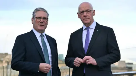 Sir Keir Starmer and John Swinney. Sir Keir has grey hair and Swinney has a bald head. They are both wearing glasses and dark suits with white shirts. Sir Keir is wearing a blue tie and Swinney a purple tie.