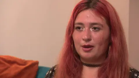 Stella sits on a sofa and speaks off camera. She has dyed red hair, hooped gold earrings and a black choker necklace.