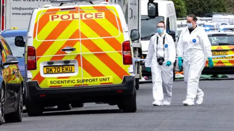 Two forensic officers in surgical masks and full-body white suits walk to liveried police van near the attack scene