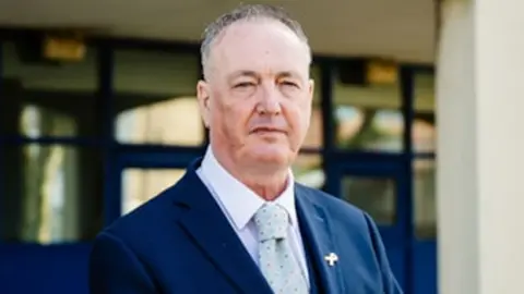 Lancashire PCC Clive Grunshaw standing outside a building with blue-framed windows. He has short grey hair and is wearing a navy suit jacket, white shirt and pale blue patterned tie