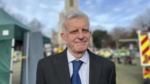 George Carden/BBC Trevor muten wearing a suit jacket and shirt and tie standing infront of a large church with emergency service vehicles behind him
