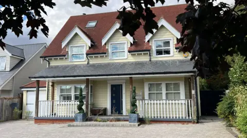 John Fairhall/BBC The front of a light yellow weatherboarded house of three storeys. It has a large driveway and two potted plants by the front door.