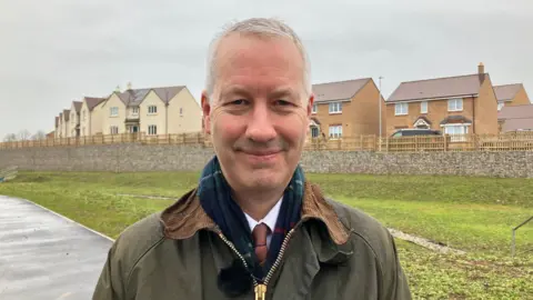 BBC Gideon Amos smiling at the camera in front of a new row of houses on a new housing estate in Taunton