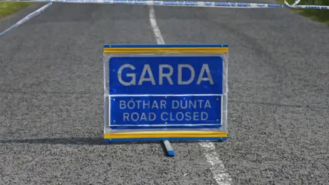 A blue sign placed on a road with white markings. The sign says Garda and road closed in both Irish and English. 