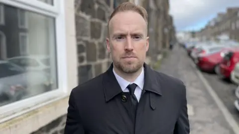 Detective Inspector Forbes Wilson is a young, bearded man with reddish hair. He is wearing a black raincoat, white shirt and black tie and is standing in a street of stone buildings with a line of cars parked facing towards or away from the kerb.