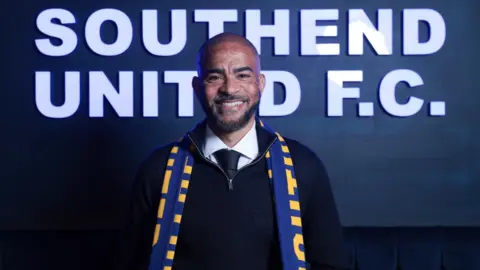 Kieron Dyer stands in front of a dark blue sign with white writing on it saying Southend United FC. He is wearing a zip-neck jumper with white shirt and dark tie underneath, with a blue and yellow scarf around his neck