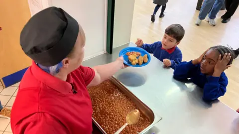 A dinner lady serves beans over a metal counter onto the plate held by a child. 