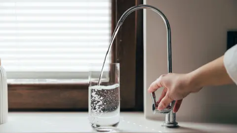 A hand turning a kitchen tap from which water is flowing into a glass