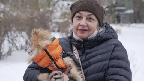 Olena stands holding her dog wearing a black puffer jacket and brown woolly hat with snow in the background.