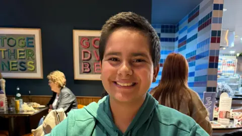 Family handout A boy smiles widely at the camera, he is wearing a green hoodie. He is sitting in a restaurant and other customers can be seen sitting on tables behind him