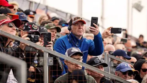 IOM TT A crowd sitting on a grandstand, which sees men wearing TT caps. One wearing a bright blue jacket is standing up filming on his phone.