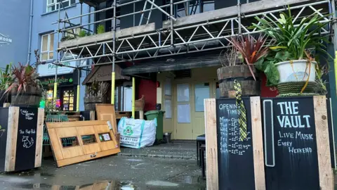 Alex Osborne/BBC Image shows ex-bar The Vault, with closed doors, old furniture and plants lying outside and scaffolding overhead. Signs still display showing opening times and 'live music every weekend.' 