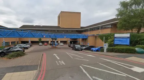 Google A blue and white hospital sign on the right is near bushes. A road near a building includes the word "In" on the ground in white along with an arrow. There is also an arrow for vehicles going in the opposite direction.