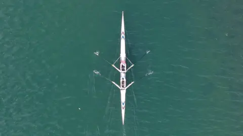 Peterborough From Above A white rowing boat with two rowers gliding through the water.