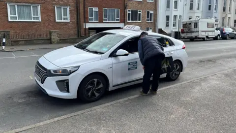 BBC A white taxi waits at a rank in Seaford. An older man is speaking to the driver through the front passenger window.