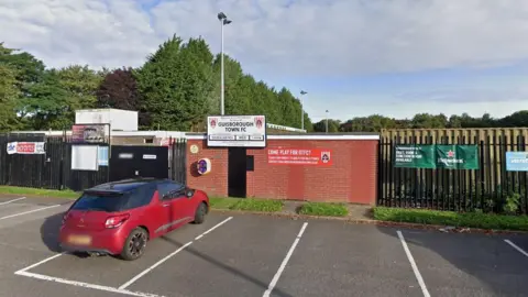 The outside entrance of Guisborough Town FC. The gates are closed and the old red brick turnstile building is locked up. There is a large Guisborough Town FC sign on the building, with the top of the main stand behind. In the distance are large green trees and a blue sky. In the foreground is a parked red car.