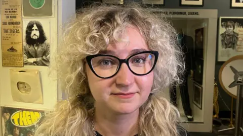 A woman with long, blonde, curly hair. She is wearing black glasses. 
