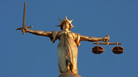 A gold-coloured statue of Lady Justice, a woman wearing a crown and holding scales and a sword in her hands, with her arms outstretched. 