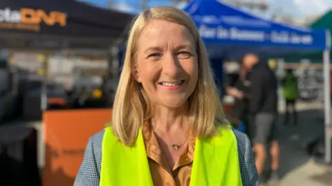 BBC/George Carden A woman with shoulder-length blonde hair smiles, staring directly into the camera. She is wearing a high-vis jacket and is stood on front of stalls at the workshop.