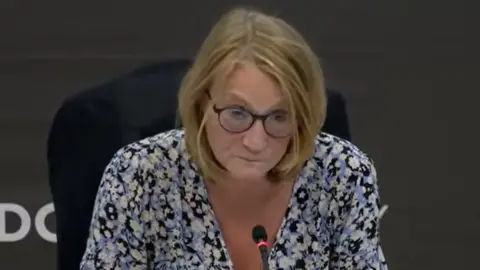 London Assembly Lyn Garner is seen with blonde shoulder-length hair and glasses wearing a floral top, looking downward towards a microphone at a committee hearing desk. 