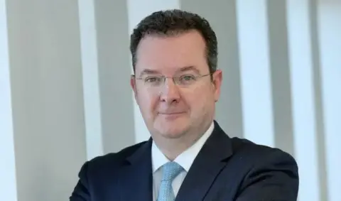 A man short curly hair is smiling toward the camera. He is wearing rimless glasses, a dark suit jacket, white shirt and blue tie. 