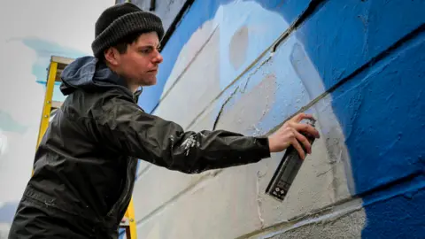 Angry Dan Angry Dan wearing a jacket and woollen hat spraying paint on a wall