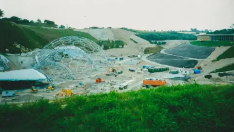 Eden Project The Eden Project during construction. The biomes are frames with just a couple of cells covered in plastic. The clay pit is bare and there are metal containers spread around the basin. 