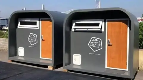 Amazing Grace Spaces Two grey housing pods with a brown wooden door and a curved roof. There is an Amazing Grace logo on the right. both pods have a window above the logos. 