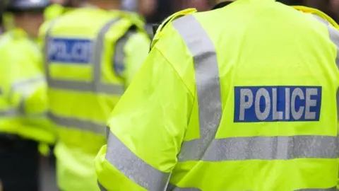 Getty Images Three police officers standing outside with their backs to the camera. They are all wearing yellow fluorescent jackets which have silver stripes on the back and arms and writing which says POLICE on the back. 