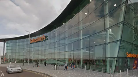 External glass and steel front of the six-storey Sainsbury's store against the backdrop of a grey sky.