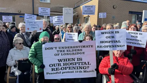 An image of elderly protestors outside Longridge Hospital. They are holding placards saying "when is our hospital reopening."