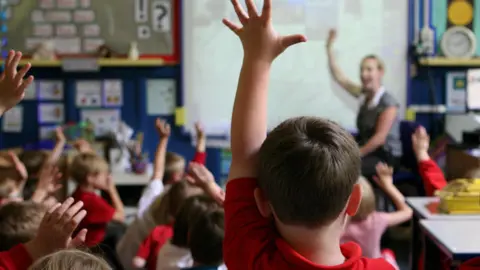 PA Media A white boy with dark hair wearing a red shirt raises his hand in a primary school class