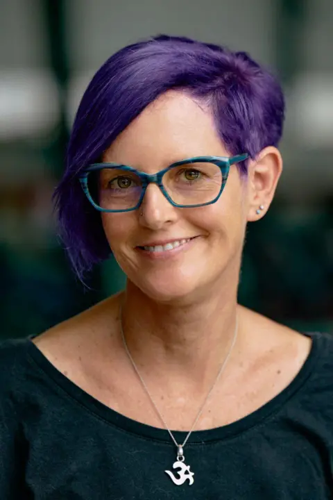 Chloe Orkin Head-and-shoulders portrait of a woman with short purple hair and blue glasses, wearing a dark top and a pendant necklace.