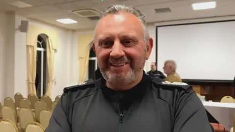 BBC middle aged man with grey hair and a black shirt he is in a room with rows of chairs behind him.