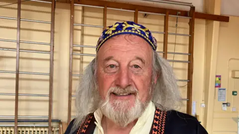 A man with long grey hair and a beard in costume. He has a dark coloured robe on and a blue and yellow hat