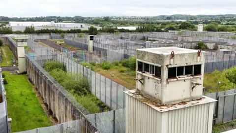 PA Media The former Maze prison near Lisburn, a large, grey building overgrown with weeds, there is a white tower in foreground