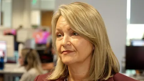 Alison honour is wearing a red blazer and looking to her right out of frame, in the background blurred call handlers and screens can be seen