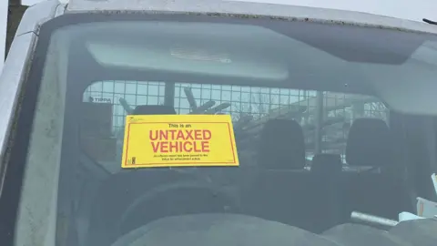 Cambridgeshire Police A close up of a vehicle windscreen. It has a bright yellow sticker above its steering wheel. It says This is an untaxed vehicle in black and red lettering