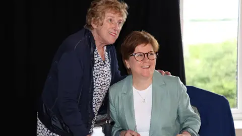 PA Media smiling nicola sturgeon in a green jacket and white top is sitting next to a standing woman in blue jacket after signing her book