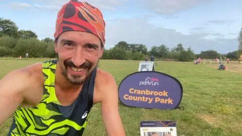 Adam Holland smiling and wearing a yellow and black running vest and red bandana holding a Parkrun barcode in front of a Cranbrook Country Park sign in a field.