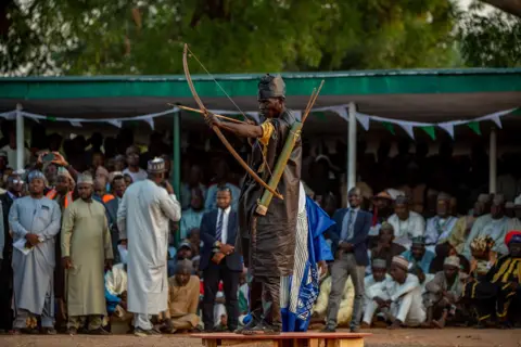 Sani Maikatanga An archer in traditional clothes readies to fire his arrow. A crowd of onlookers can be seen behind him.