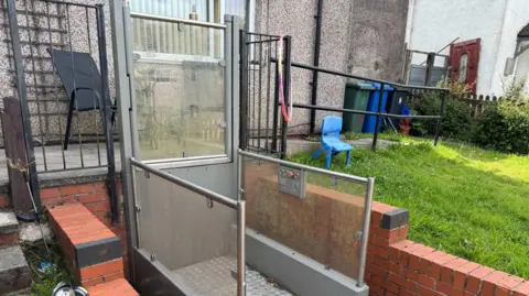An image of an outside lift, it is encased in a red bricked wall and is in the front garden of Mulcahy's house in Bacup