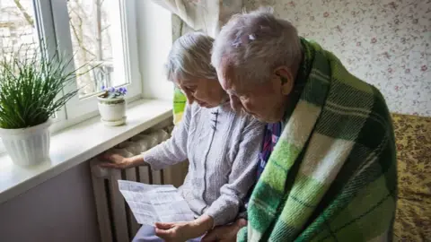 An older couple looking at a letter. The woman has her hand on the radiator and the man is wearing a blanket around his shoulders.