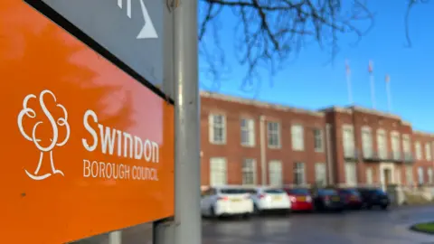 Close-up of Swindon Borough Council logo on a sign with the red brick HQ blurred in the background.