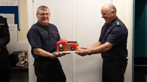 Keith Prince Keith Prince being handed a cake in the shape of a fire engine on his retirement evening. He is smiling with a colleague of his who is giving him the cake.