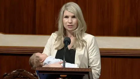 Brittany Pettersen with long blonde hair wearing a white jacket and black top cradling her baby wearing blue