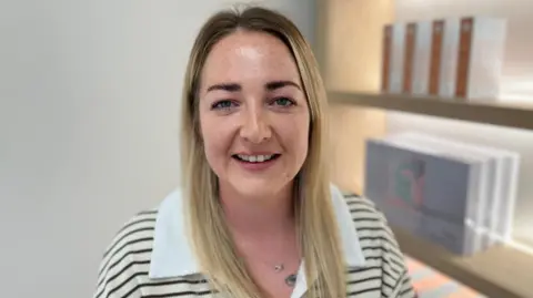Hannah has long, straight blonde hair and is smiling at the camera. She is wearing a cream and black striped top, with a white collar. She is stood next to shelves displaying products in a clinic. It is a head and shoulders shot.